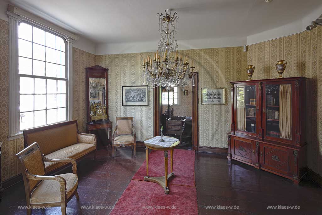 Remscheid Hasten, Haus Cleff, historisches Zentrum, Dauerausstellung in demEmpire Zimmer, mit Holzboden und einem langen roten Teppich, welches ausgestattet ist mit antiken Moebeln wie einem Glasvitrinenschrank mit Bchern und zwei Vasen, Bildern, einen Tuerrahmen, einer Spiegeluhr, zwei Sesseln, einem Sofa vor einem grossen Fenster und einem Tischchen unter einem Kronleuchter ; Remscheid-Hasten, House Cleff, historcal centre, permanent exhibition in a empire living room with antiquely furniture