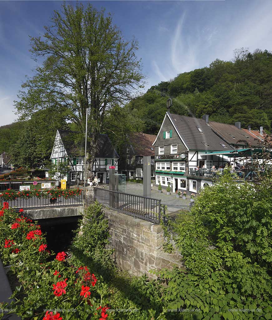 Solingen Unterburg, Schlossbergstrasse, bluehende Geranien im Vordergrund von einem Kopsteinpflasterplatz mit Briefkasten der Deutschen Post AG, Burger Brezelbaecker Denkmal, Stadtsparkasse Solingen, eine von zwei alteren Damen benutzte Bank  und Cafe Meyer, Fachwerk- und Schieferhauser, Brezel auf dem Dach von dem Caf; Solingen-Unterburg, Schlossbergstrasse,with blooming geranium in front of a place of cobble stone pavement, with a german letter box, Burger pretzel baker historical monument, cafe Meyer, with frame houses and houses of shist, a pretzel is on the roof of the caf