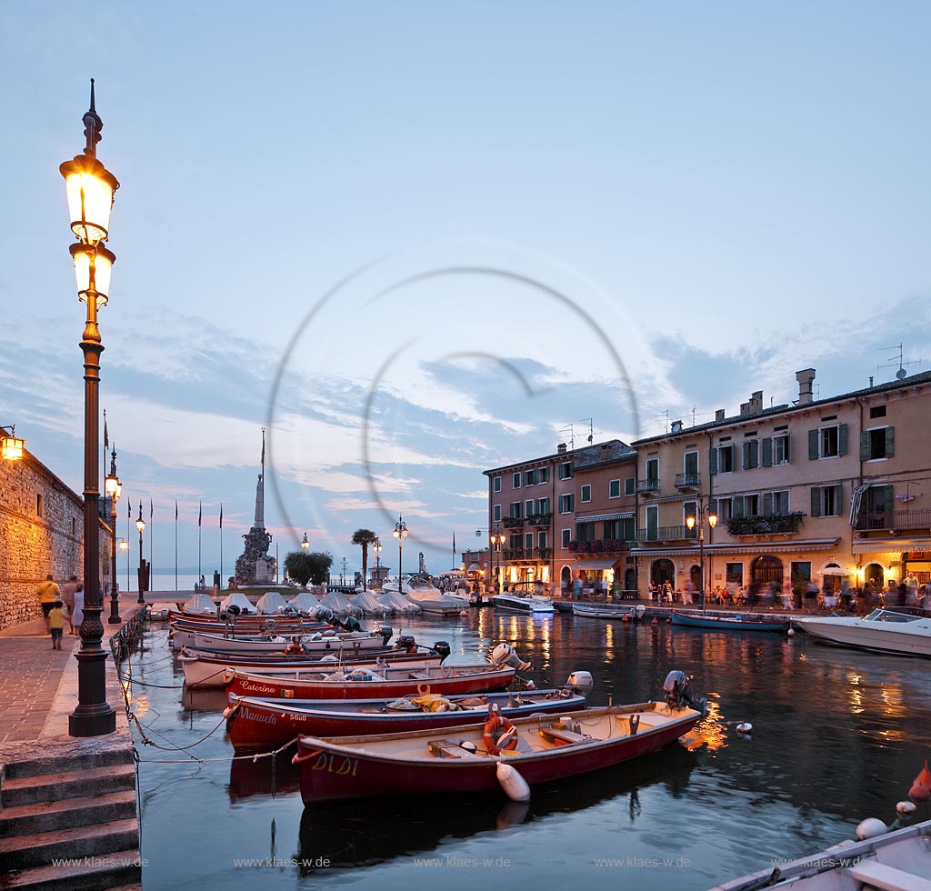 Lazise, Blick ueber den Hafen Richtung Gardasee mit bunten Fischerbooten und Haeusern sowie dem Denkmal fuer Kriegsgefallene zur blauen Stunde; Lazise, view over the port with colourful fishing boats, houses and war victim memoirial during blue hour