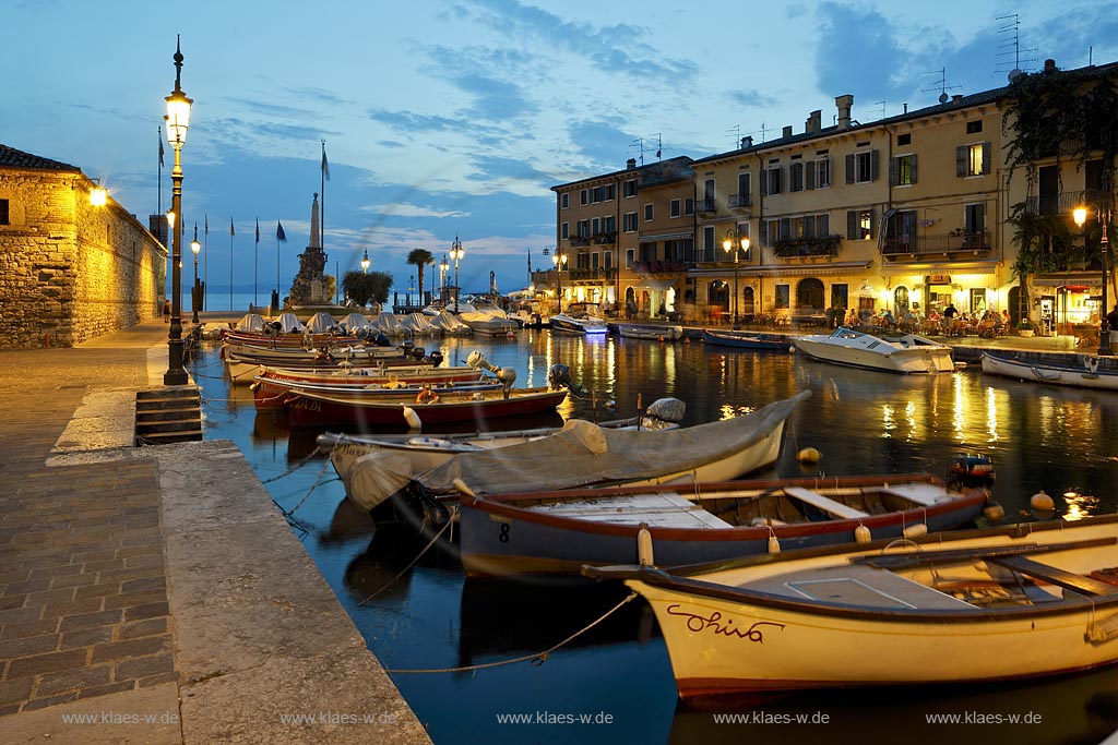Lazise, Blick ueber den Hafen Richtung Gardasee mit bunten Fischerbooten und Haeusern sowie dem Denkmal fuer Kriegsgefallene zur blauen Stunde; Lazise, view over the port with colourful fishing boats, houses and war victim memoirial during blue hour