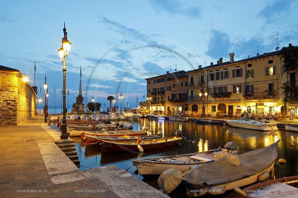Lazise, Blick ueber den Hafen Richtung Gardasee mit bunten Fischerbooten und Haeusern sowie dem Denkmal fuer Kriegsgefallene zur blauen Stunde; Lazise, view over the port with colourful fishing boats, houses and war victim memoirial during blue hour