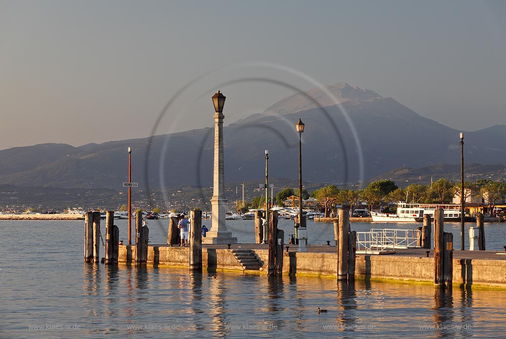 Peschiera, Abenstimmung mit Blick ueber die Hafenmole mit Leuchtmast zum Gardasee im stimmungsvollen Abendlicht der untergehenden Sonne, im Hintergrund der Monte Baldo; Peschiera view over the mole with lightmast in evening sunset light, in the background mountain of Monte Baldo