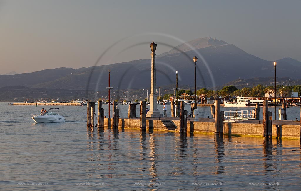 Peschiera, Abenstimmung mit Blick ueber die Hafenmole mit Leuchtmast und Motorboot zum Gardasee im stimmungsvollen Abendlicht der untergehenden Sonne, im Hintergrund der Monte Baldo; Peschiera view over the mole with lightmast an motorboat on the lake  in evening sunset light, in the background mountain of Monte Baldo