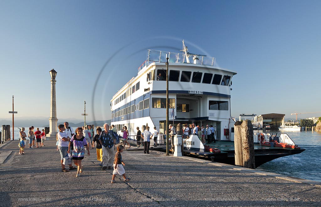 Peschiera, Abenstimmung mit Blick ueber die Hafenmole mit Leuchtmast und Menschen, anlegendes Faehrschiff Brescia mit Passagieren an Bord im stimmungsvollen Abendlicht; Peschiera view over the mole with lightmast and humans, waiting for the landing ferry ship Brescia in evening light