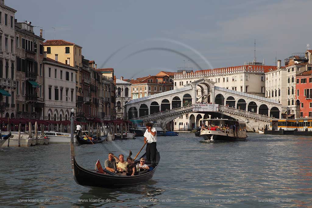 Venedig, Rialto, Blick von Riva del Carbon auf Kanal Grande mit der Rialtobruecke Ponte di Rialto, Gondeln mit Gondoliere einer telefonierend mit Handy, Mobiltelefon mit T Shirt weiss und zwei Paare in Gondel sitzend; Venice Rialto view from Riva del Carbon to Canal Grande with Rialto bridge, gondola and gondolier one is talking on the mobile phone in white t-shirt, two couple are sitting in gondola