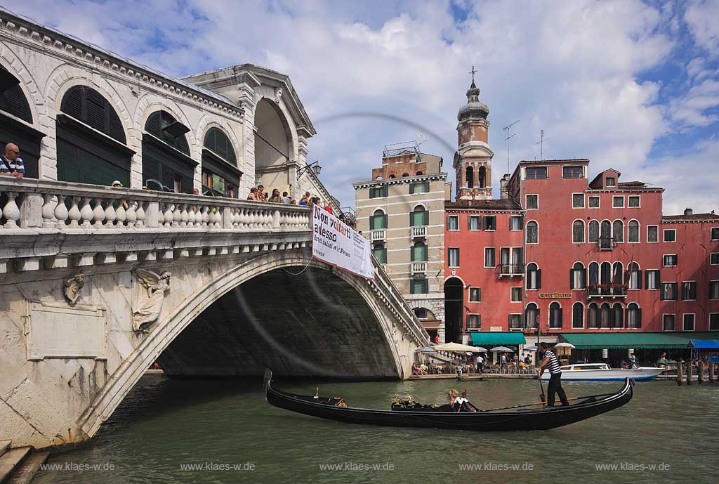 Venedig Bruecke Ponte di Rialto mit Kanal Grande seitlich fotografiert mit darunter herfahrender Gondel mit Gondoliere und jungem Paar, huebsche junge Frau in Gondel sitzend, Menschen auf Bruecke stehend, gegenueber Riva del Ferro mit dem Kirchturm Campanile der Kirche Chiesa di San Bartolomeo; Venice sideways view to bridge of Rialo with Canal Grande, a gondola with gondolier and a young couple, a pretty young woman sitting inside of gondola in the background Riva del ferro with the tower of church of San Bartolomeo