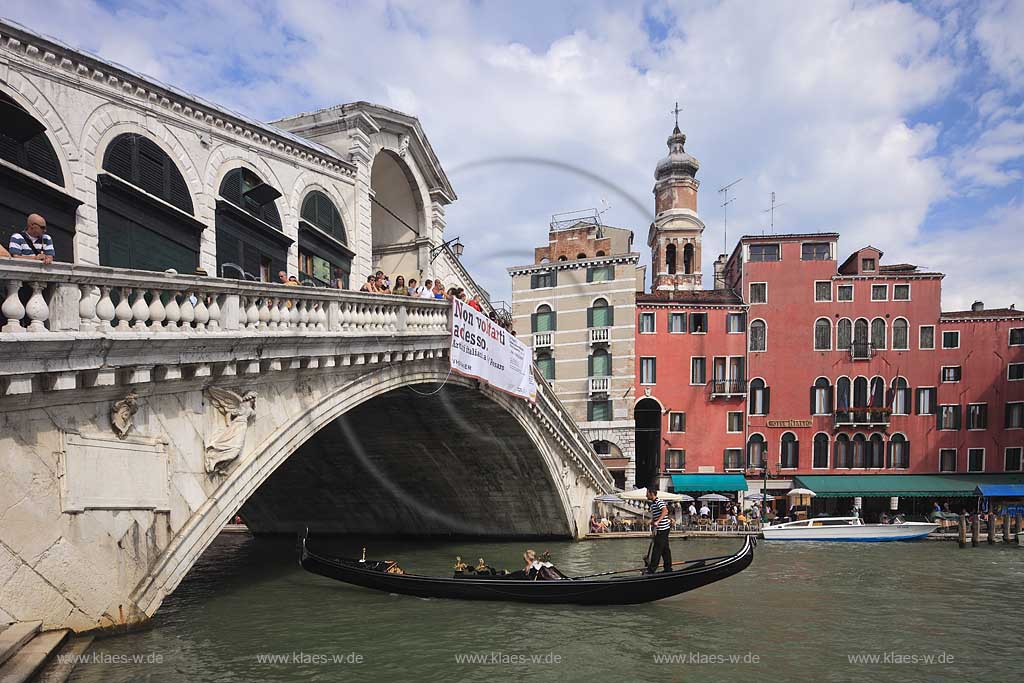 Venedig Bruecke Ponte di Rialto mit Kanal Grande seitlich fotografiert mit darunter herfahrender Gondel mit Gondoliere und jungem Paar, huebsche junge Frau in Gondel sitzend, Menschen auf Bruecke stehend, gegenueber Riva del Ferro mit dem Kirchturm Campanile der Kirche Chiesa di San Bartolomeo; Venice sideways view to bridge of Rialo with Canal Grande, a gondola with gondolier and a young couple, a pretty young woman sitting inside of gondola in the background Riva del ferro with the tower of church of San Bartolomeo
