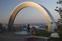 Kiew Blick vom Vladimirskaya Gorka Park durch den Bogen der Voelkerfreundschaft zwischen der Ukraine und Russland auf den Fluss Dnepr sowie die Hochhaeuser  am Westufer im roetlichen Licht der untergehenden Sonne . The friendship Arch: sculptor A. Skoblikov and Architect I. Ivanov and others. Dedicated to the unification of Russia and Ukraine, but called "The Yoke" by kievans. The rainbow shape arch is 50 metres in diameter. Viewing deck were most of the east bank can be viewed, Troeschina and towards the north of city. Podil and Obolon. Undemeath the Friendship Arch are two statues. The left one is made from bronze and depicts a Russian and Ukrainian worker holding up the Soviet Order of Friendship of People the other one is made from granite and depicts the particants of the Pereyaslavska Rada of 1654.