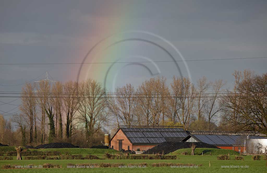 Kleve Niederrheinlandschaft mit Gehoeft mit Kollektoren, Solarenergie auf den Daechern,Strommasten und Stomleitungen,schwarzerHimmel, Sonnenschein und Regenbogen, kahle Baeume im Fruehling; Klever Netherrhine landscape with 