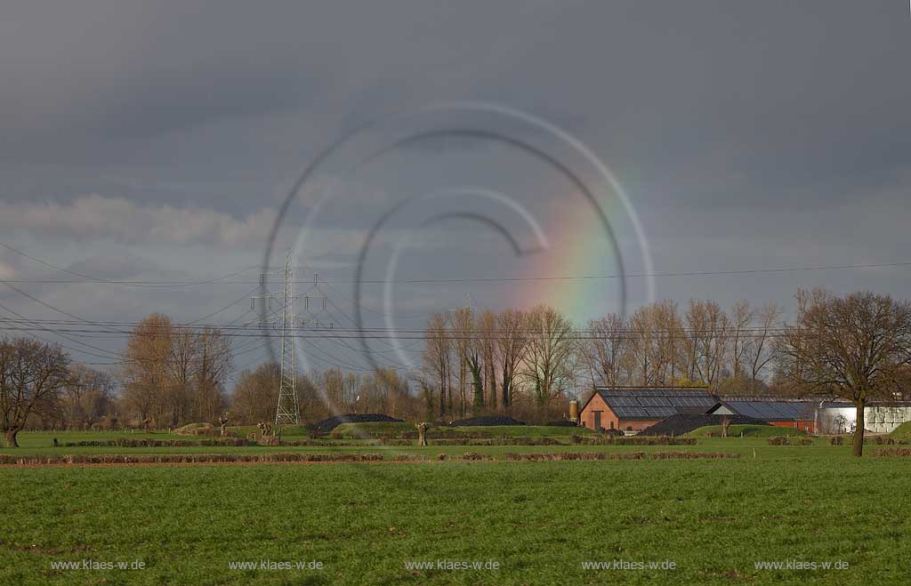 Kleve Niederrheinlandschaft mit Gehoeft mit Kollektoren, Solarenergie auf den Daechern,Strommasten und Stomleitungen,schwarzerHimmel, Sonnenschein und Regenbogen, kahle Baeume im Fruehling; Klever Netherrhine landscape with 